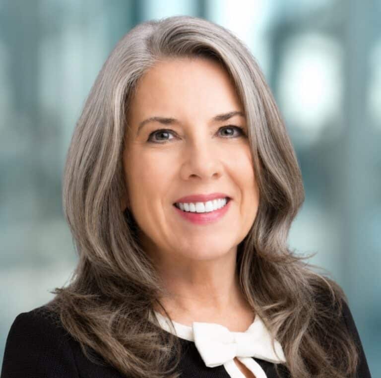 Smiling woman with long gray hair wearing a black jacket and a white blouse with a bow, standing in front of a blurred indoor background.