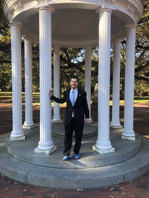 A person in a suit and blue shoes stands by a white, columned gazebo outdoors, surrounded by trees and sunlight, as Stewart Banner attends the grand opening of UNC Kenan-Flagler’s new business school building.