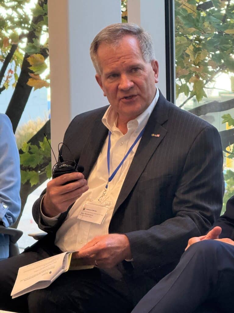 A man in a dark suit and white shirt sits by a window holding a microphone and papers, with a lanyard and name badge around his neck at the Nordic Business Breakfast. Trees with autumn leaves are visible outside, setting the scene as he discusses trends in transatlantic trade.
