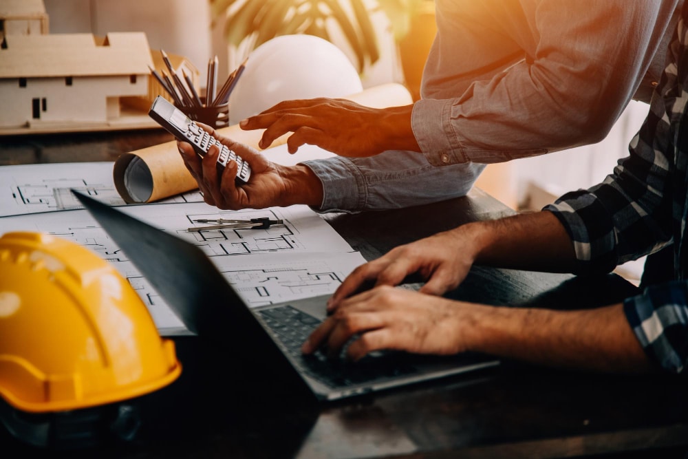 Two people work at a desk with building plans, a laptop, and a calculator. A yellow hard hat and architectural tools are nearby, suggesting a construction or engineering project.