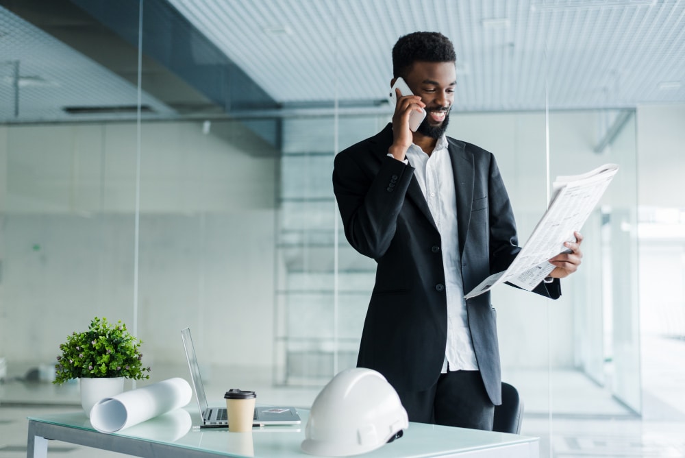 A man in a suit stands at a desk, smiling while talking on the phone and holding architectural plans. The desk has a hard hat, plant, laptop, and coffee cup. The background is a modern, glass-walled office.