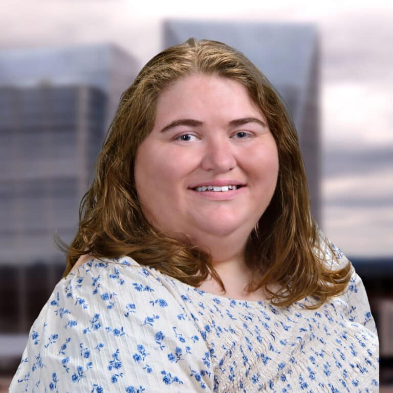 A smiling person with long, light brown hair wearing a white top with blue flowers, standing in front of blurred city buildings and a cloudy sky.