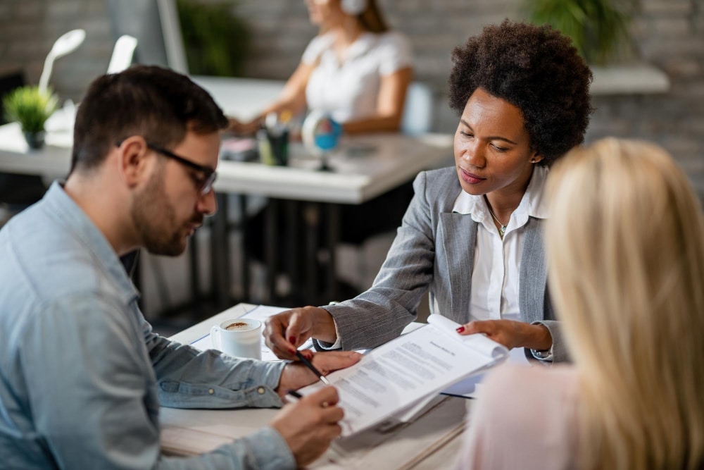 A commercial real estate attorney in Atlanta reviews documents with a man and a woman at a desk in an office, while another woman works at a computer in the background.