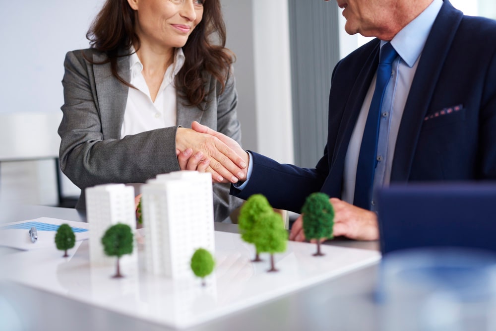 Two people in business attire shaking hands over a table with a scale model of buildings and trees, indicating a successful architectural deal facilitated by a commercial real estate attorney in Atlanta.