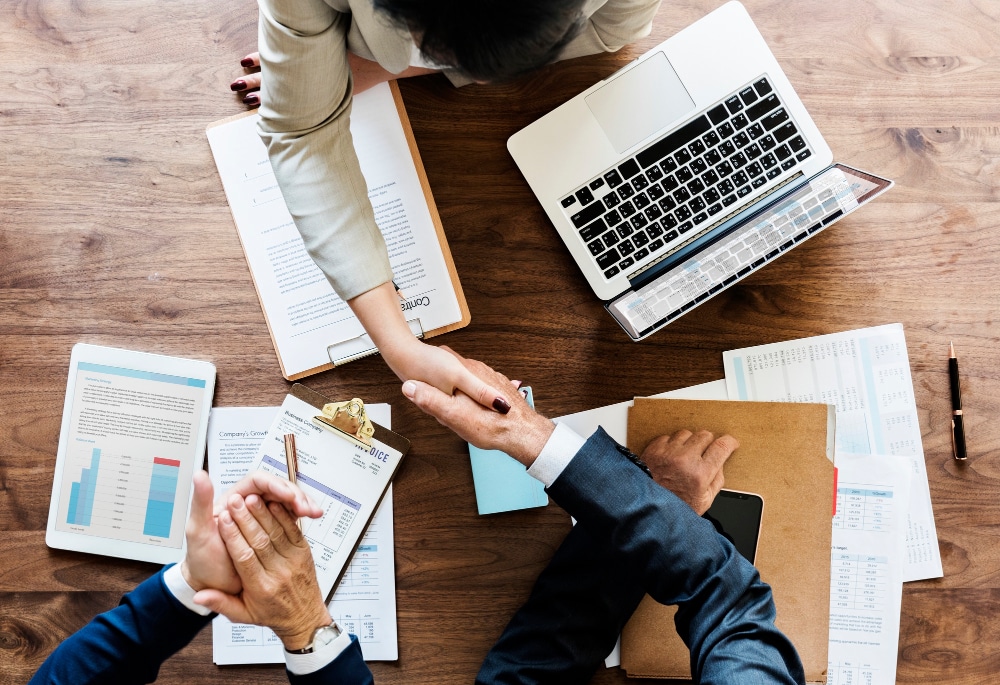 Three people at a wooden table with documents, a clipboard, and a laptop, shaking hands, suggesting a business meeting or agreement.