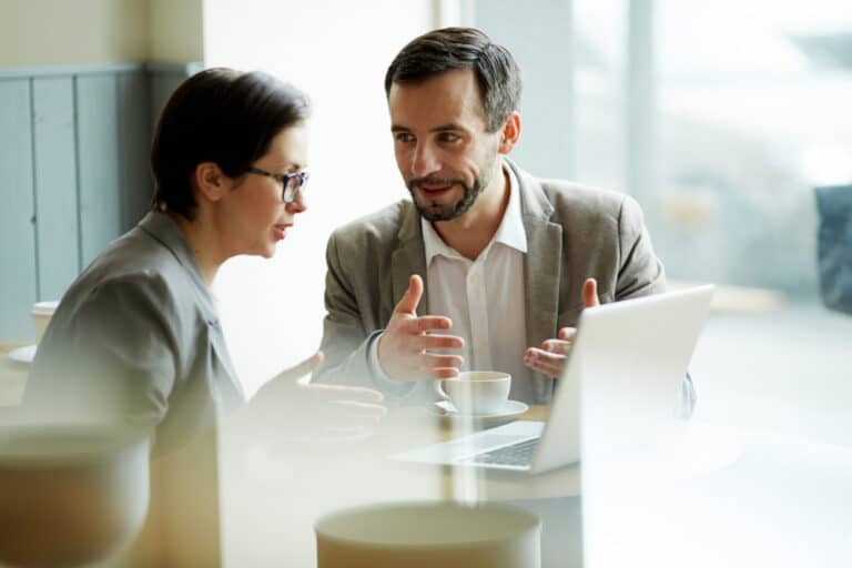 Two people in business attire sit at a table with a laptop and coffee cups, having a focused conversation in a modern, bright cafe or office space.