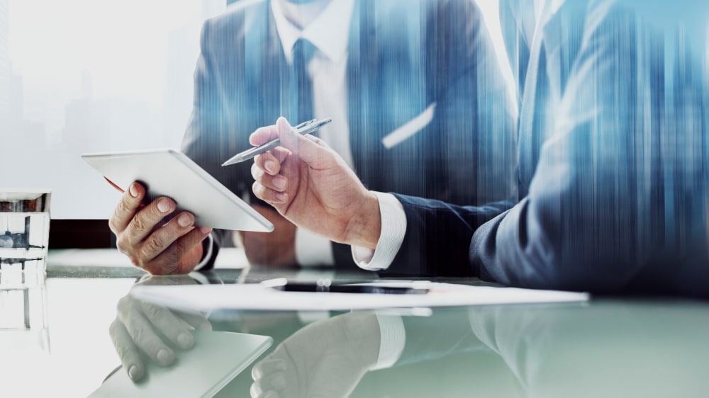 Two people in business suits sit at a glass table, one holding a tablet and the other gesturing with a pen, suggesting a discussion or meeting.