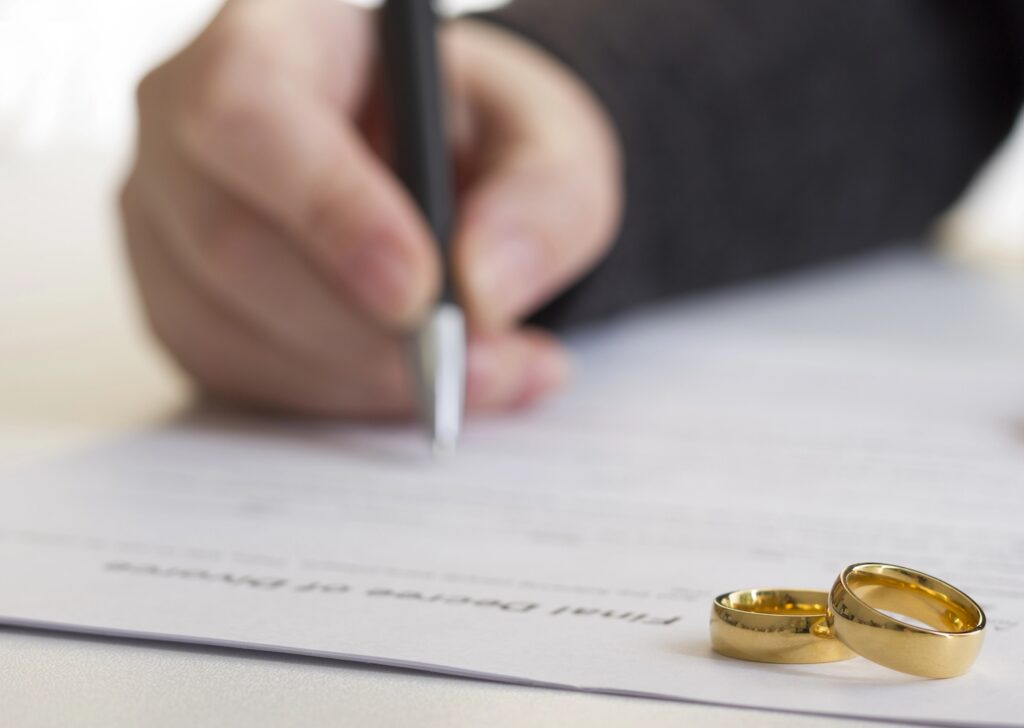 A hand signs a document labeled Final Decree of Divorce with two gold wedding rings placed on the paper.