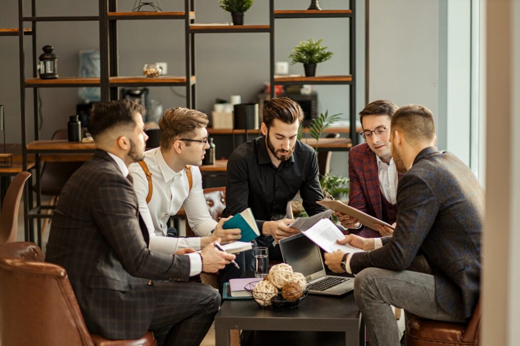 Five men in business attire sit around a coffee table in a modern office, discussing documents and holding notebooks. A laptop and decorative items are on the table, with shelves and plants in the background.