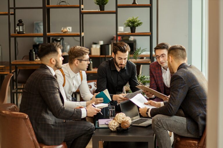 Five men in business attire sit around a coffee table in a modern office, discussing documents and holding notebooks. A laptop and decorative items are on the table, with shelves and plants in the background.