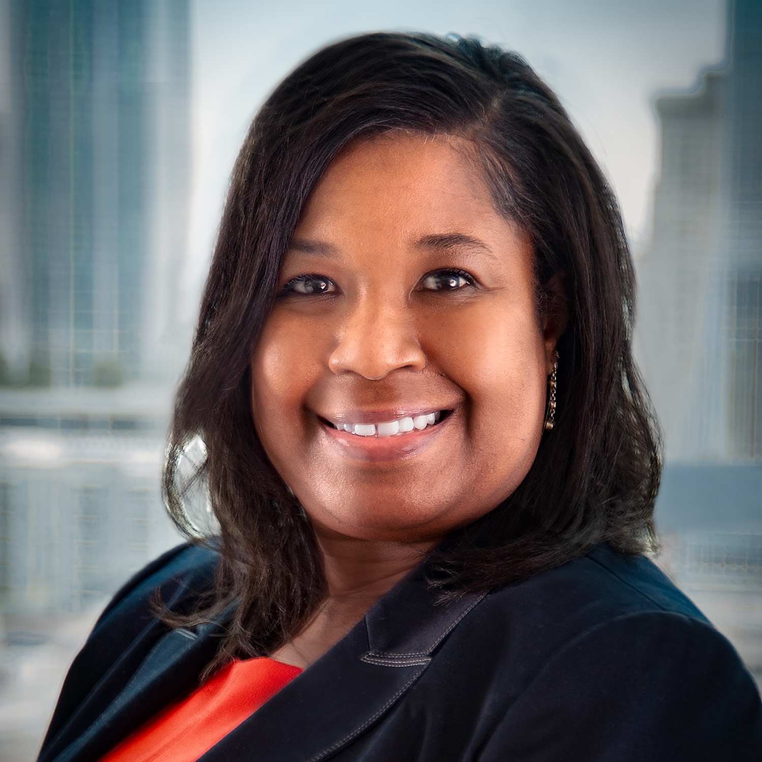A woman with shoulder-length dark hair smiles at the camera, wearing a black blazer over an orange top. A cityscape with tall buildings is blurred in the background.