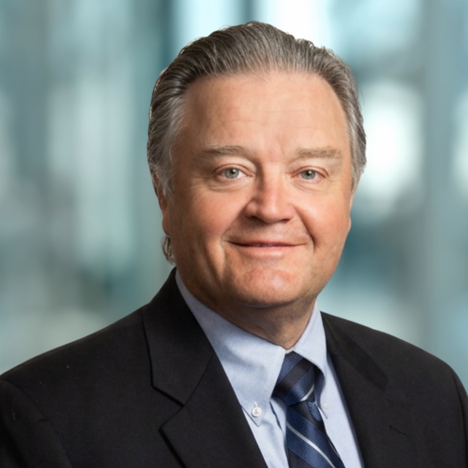 A middle-aged man in a suit and striped tie smiles at the camera with a blurred office background behind him.