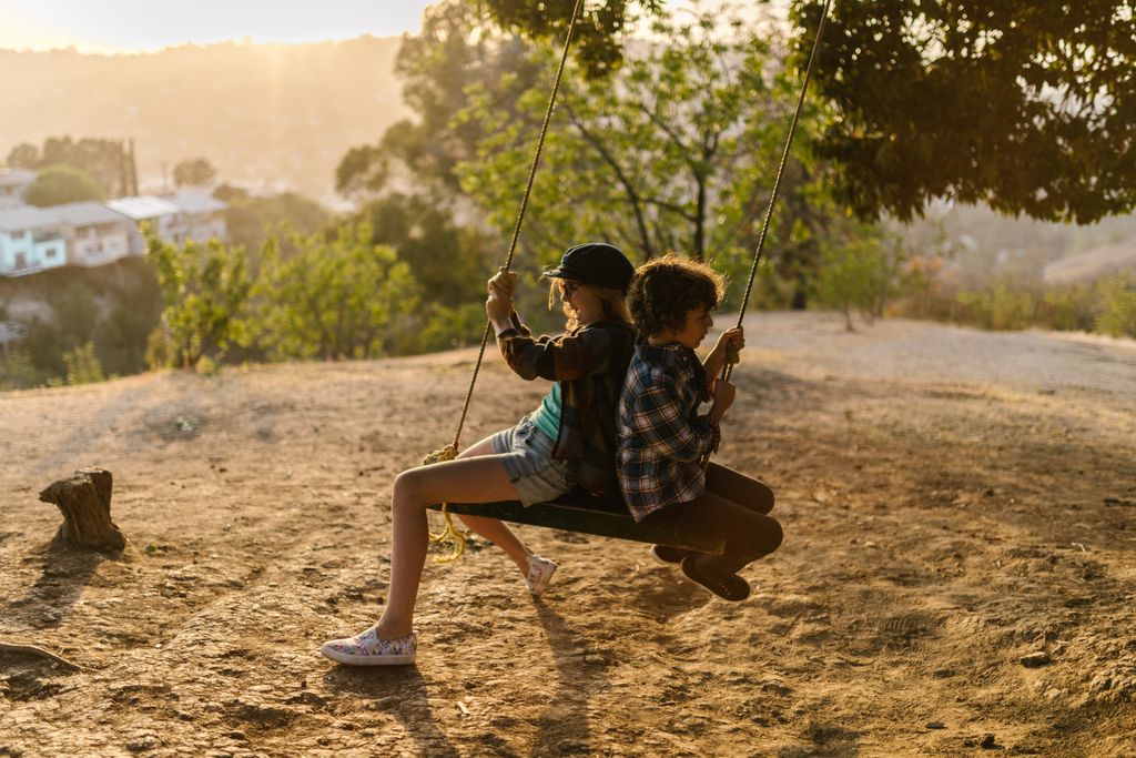 Two kids joyfully swinging together in a sunny park setting.