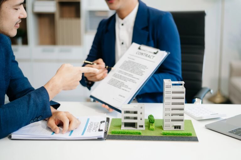 Two people in business attire review a contract at a desk featuring a small architectural model of two buildings.